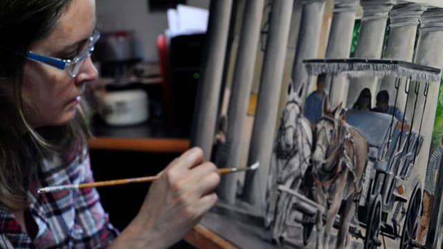 The image shows an artist painting a horse-drawn carriage with a focus on the details of the artwork being created.