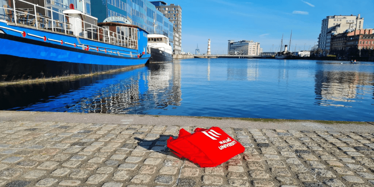 Red Malmö University tote bag displayed beside a waterfront canal in Malmö, Sweden.